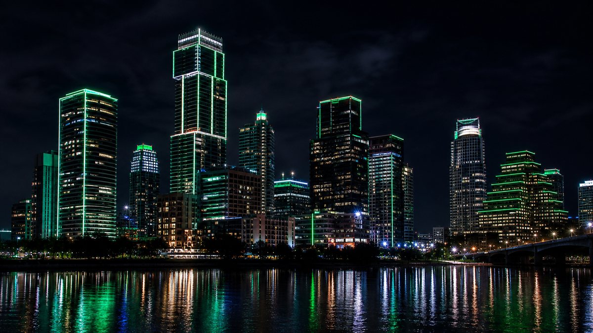 Dallas-style skyline at night with several towers lit in coordinated emerald green and white architectural LED lighting reflected in a river