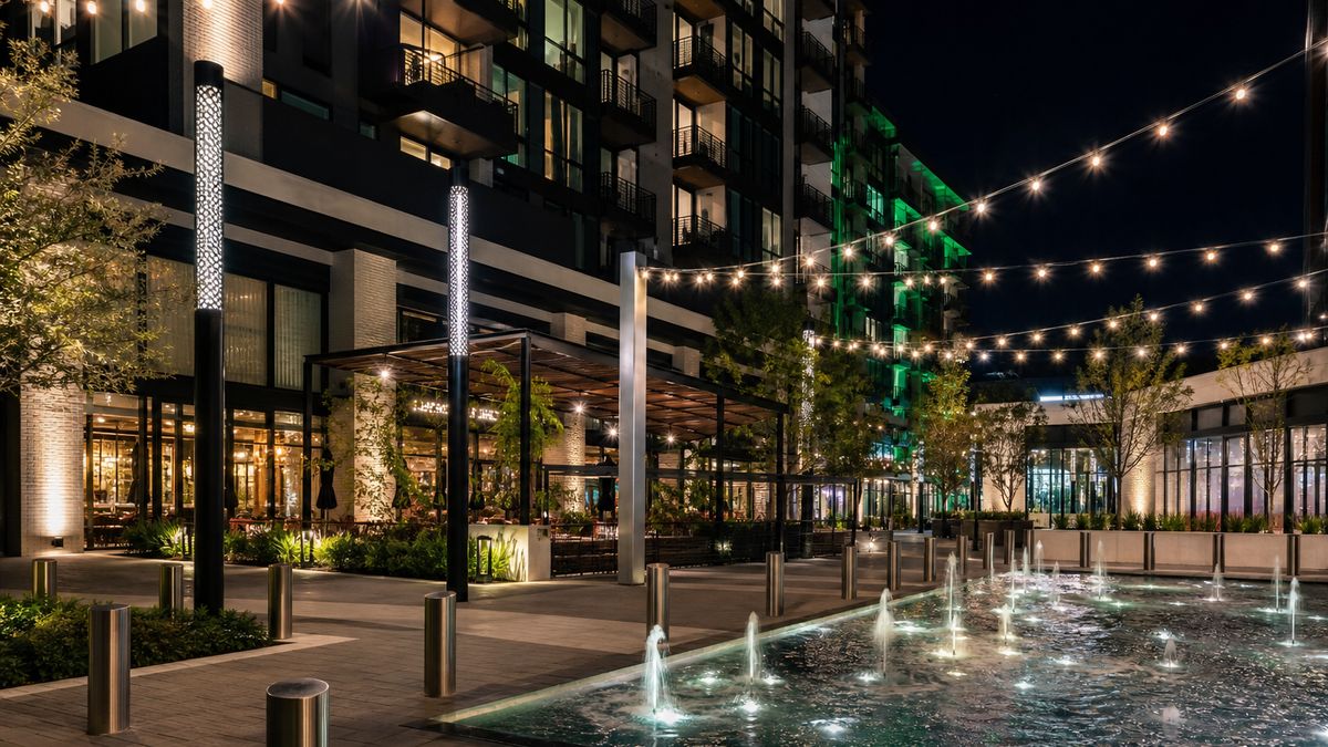 Mixed-use development plaza in Dallas at night with warm pedestrian lighting, tree uplighting, festoon lights and residential tower above retail
